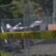 Yellow crime scene tape reading 'CRIME SCENE - DO NOT CROSS' spans a chain-link fence with a blurred outdoor background behind it.