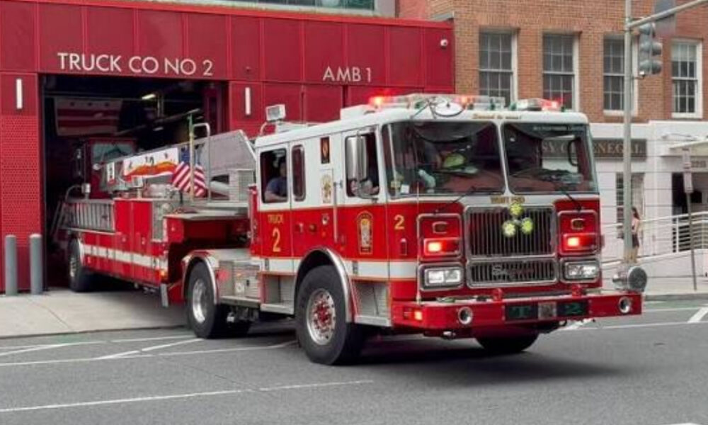Red fire engine exits a firehouse labeled 'Truck Co No 2' with emergency lights on, on a city street.