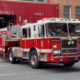 Red fire engine exits a firehouse labeled 'Truck Co No 2' with emergency lights on, on a city street.