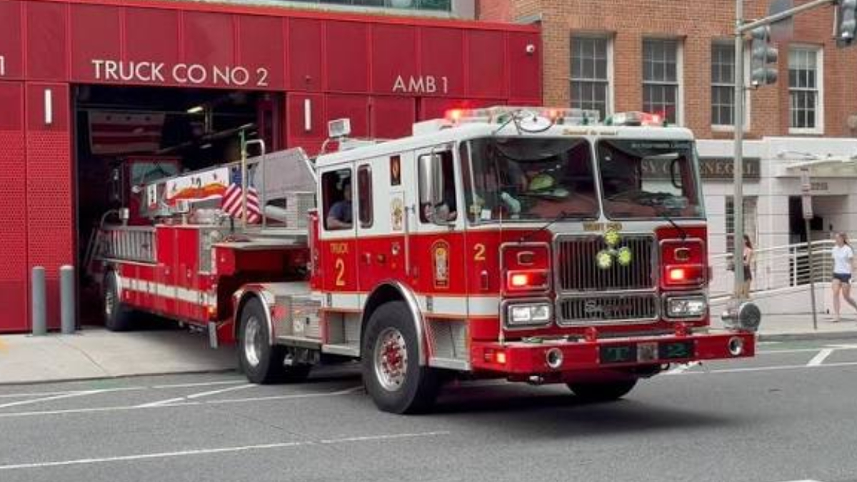 Red fire engine exits a firehouse labeled 'Truck Co No 2' with emergency lights on, on a city street.