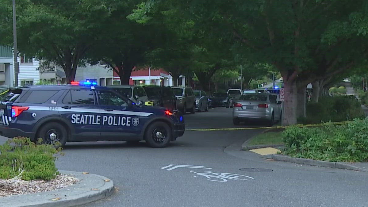 Seattle Police SUV blocks a tree-lined residential street with flashing lights and yellow tape across the road.