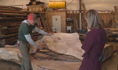 Two people in a woodworking shop examine a large, curved wooden slab with exposed grain on a work table behind them in a cluttered workshop edge.