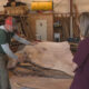 Two people in a woodworking shop examine a large, curved wooden slab with exposed grain on a work table behind them in a cluttered workshop edge.