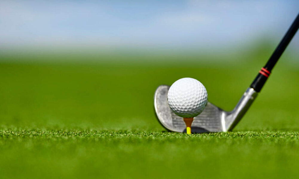 Close-up of a golf ball teed up on a yellow tee with an iron behind it on a lush green golf course.