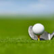 Close-up of a golf ball teed up on a yellow tee with an iron behind it on a lush green golf course.