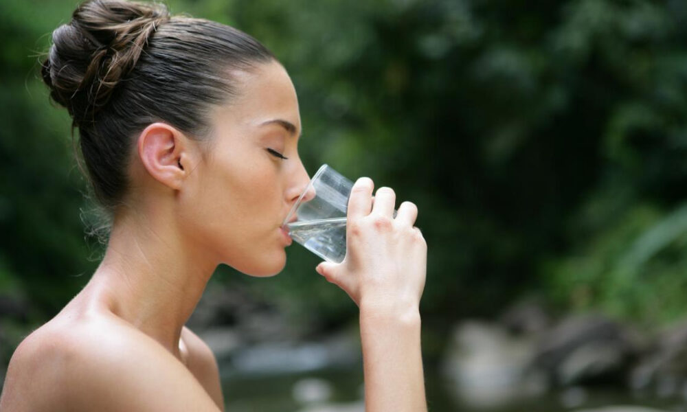 Woman in profile drinking from a clear glass of water outdoors, with greenery in the background.