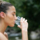 Woman in profile drinking from a clear glass of water outdoors, with greenery in the background.