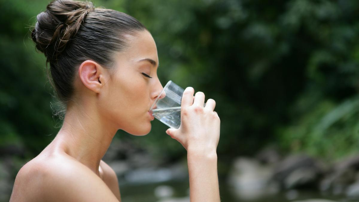 Woman in profile drinking from a clear glass of water outdoors, with greenery in the background.