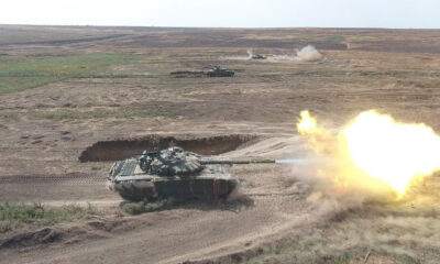 A tank fires its main gun in a dusty desert training range, with another tank visible in the distance and smoke plumes rising ahead.
