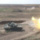 A tank fires its main gun in a dusty desert training range, with another tank visible in the distance and smoke plumes rising ahead.