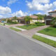 Suburban street scene with single-family homes, green lawns, and palm trees under a blue sky.