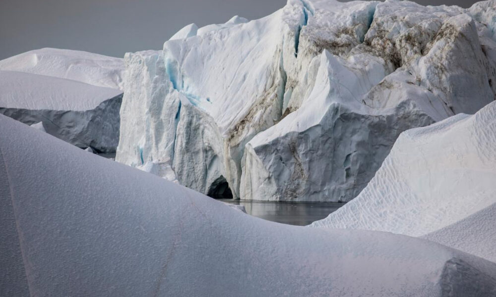Snowy glacier landscape with blue-tinged ice cliffs and a meltwater pool nearby, calm and reflective.