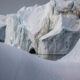 Snowy glacier landscape with blue-tinged ice cliffs and a meltwater pool nearby, calm and reflective.