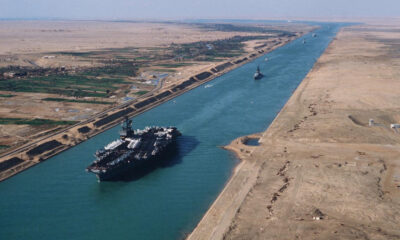Aerial view of a large container ship in the Suez Canal, with desert banks on both sides and a distant vessel.