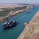 Aerial view of a large container ship in the Suez Canal, with desert banks on both sides and a distant vessel.