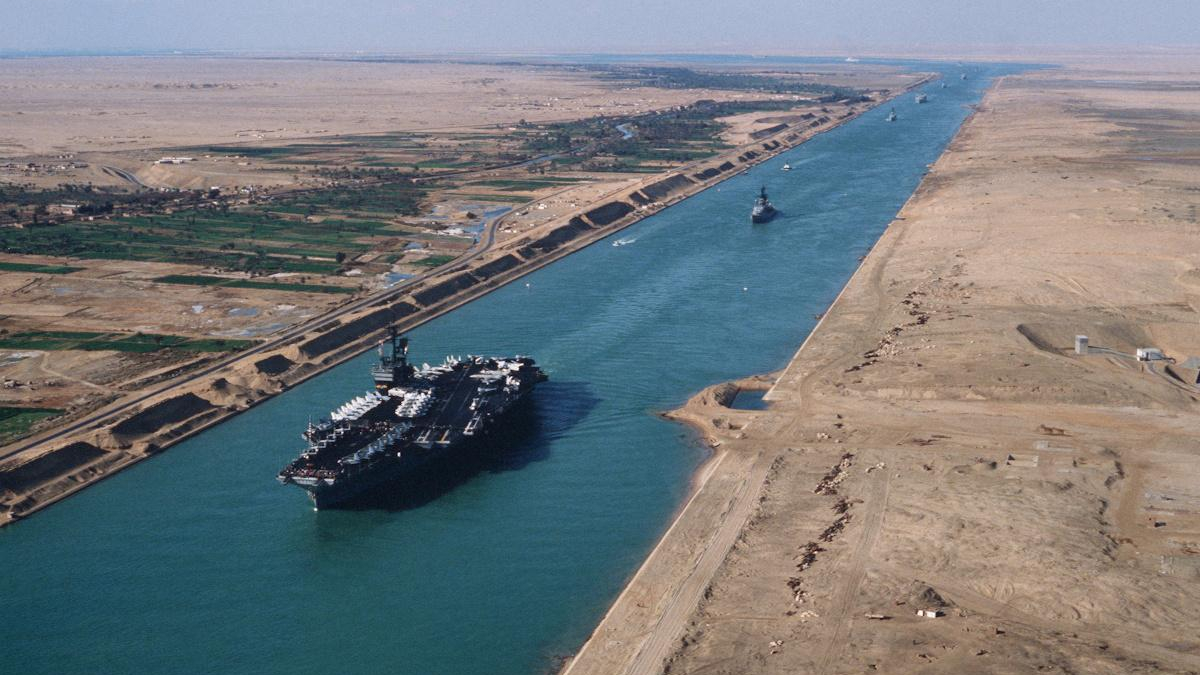 Aerial view of a large container ship in the Suez Canal, with desert banks on both sides and a distant vessel.