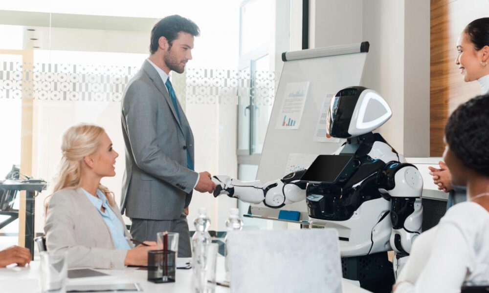 Business meeting where a robot shakes hands with a man in a gray suit, as colleagues watch in a bright office.