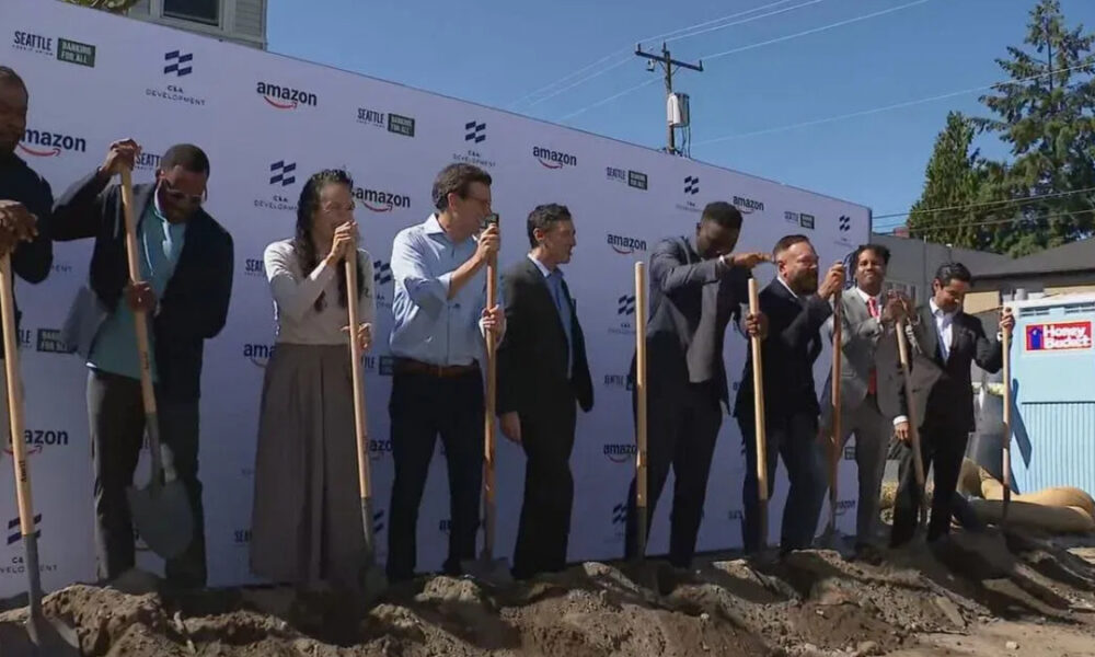 Group of professionals in business attire taking part in a groundbreaking ceremony, each holding a shovel and standing in dirt in front of a branded backdrop with logos like Amazon and Seattle Development on a sunny day.