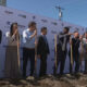 Group of professionals in business attire taking part in a groundbreaking ceremony, each holding a shovel and standing in dirt in front of a branded backdrop with logos like Amazon and Seattle Development on a sunny day.