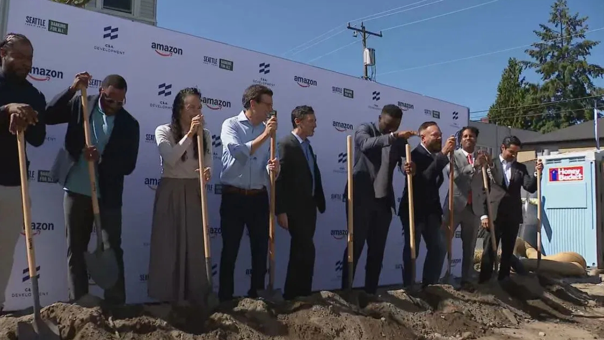 Group of professionals in business attire taking part in a groundbreaking ceremony, each holding a shovel and standing in dirt in front of a branded backdrop with logos like Amazon and Seattle Development on a sunny day.