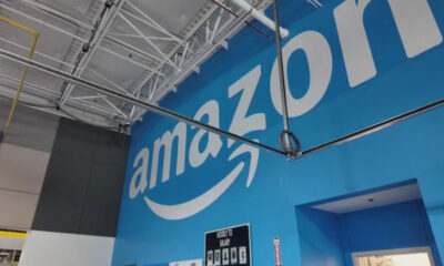 Blue wall with a large white Amazon logo inside a warehouse-like interior showing exposed ceiling pipes and wiring.