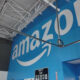 Blue wall with a large white Amazon logo inside a warehouse-like interior showing exposed ceiling pipes and wiring.