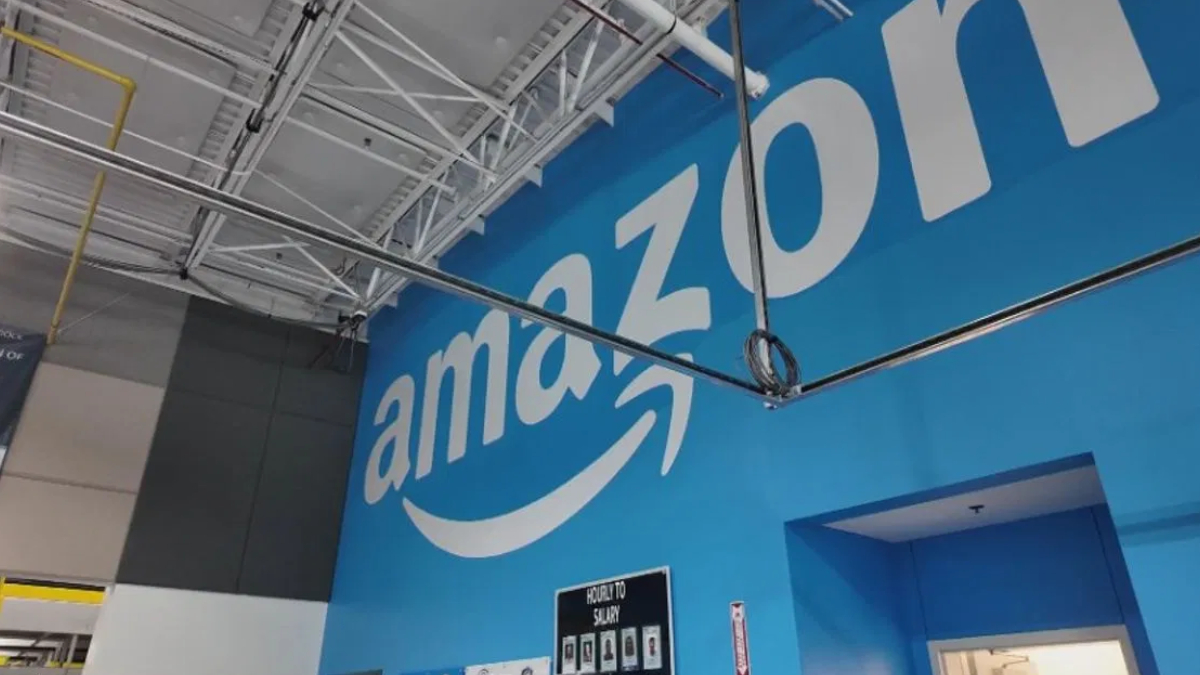 Blue wall with a large white Amazon logo inside a warehouse-like interior showing exposed ceiling pipes and wiring.