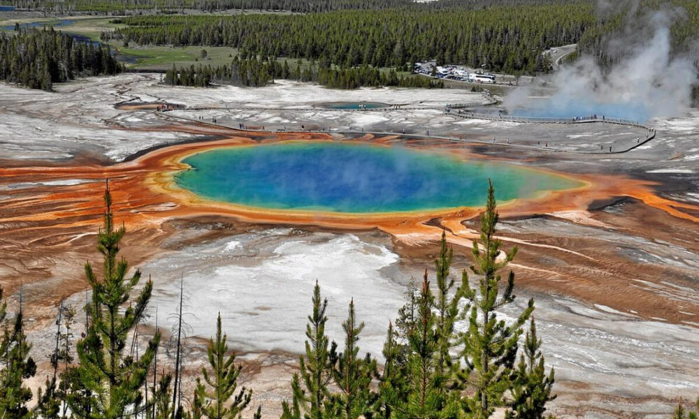 Aerial view of a vibrant blue-green hot spring with bright orange mineral rims, set in a geothermal field with forest in the background.