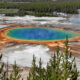 Aerial view of a vibrant blue-green hot spring with bright orange mineral rims, set in a geothermal field with forest in the background.