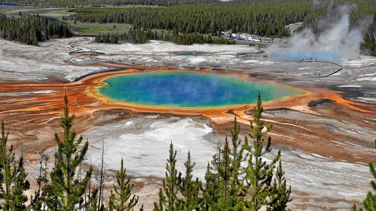 Aerial view of a vibrant blue-green hot spring with bright orange mineral rims, set in a geothermal field with forest in the background.