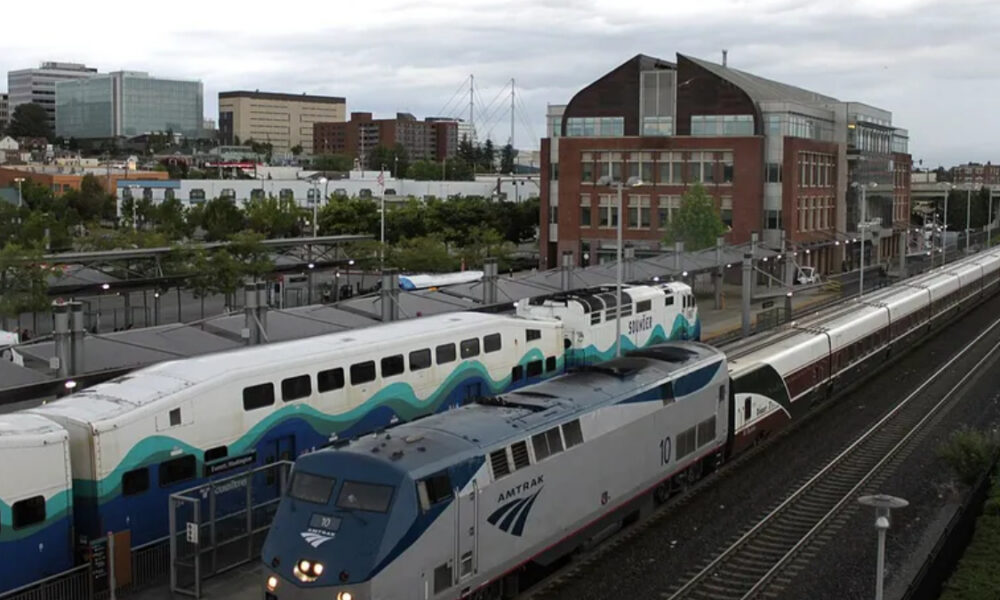 Amtrak locomotive leading a long passenger train at a city rail station, with a brick building in the background and overhead wires nearby.