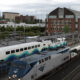 Amtrak locomotive leading a long passenger train at a city rail station, with a brick building in the background and overhead wires nearby.