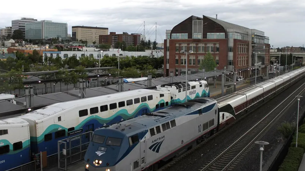 Amtrak locomotive leading a long passenger train at a city rail station, with a brick building in the background and overhead wires nearby.