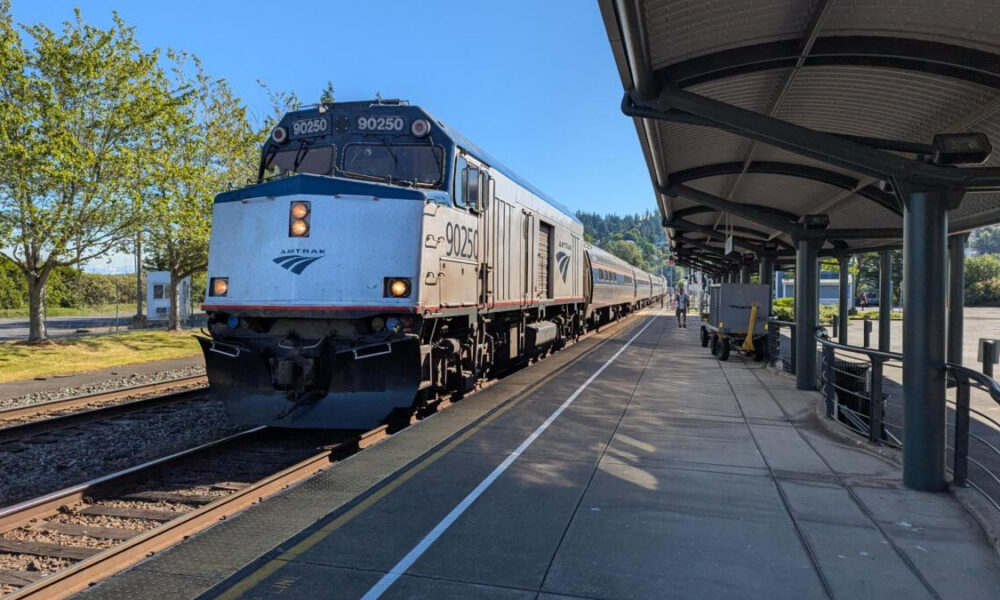 Amtrak locomotive 90250 at a sunny station platform with silver passenger cars behind it in view.