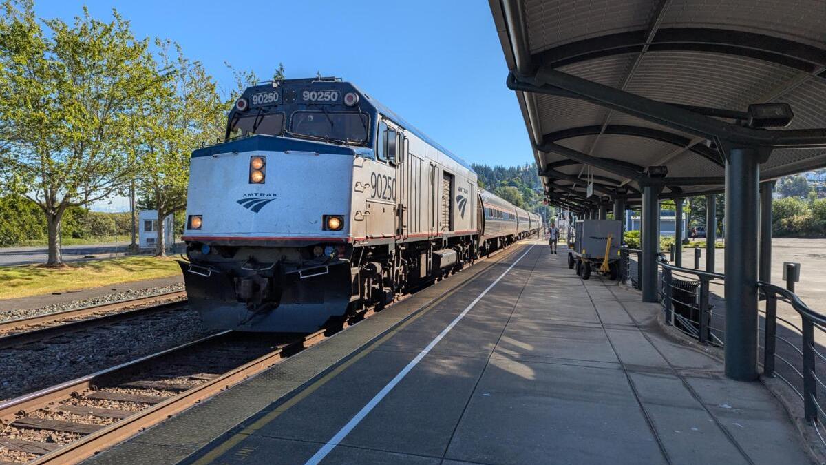 Amtrak locomotive 90250 at a sunny station platform with silver passenger cars behind it in view.