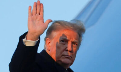 Man in a dark coat raises his right hand in a wave against a blue sky.