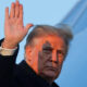 Man in a dark coat raises his right hand in a wave against a blue sky.