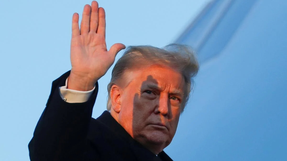 Man in a dark coat raises his right hand in a wave against a blue sky.