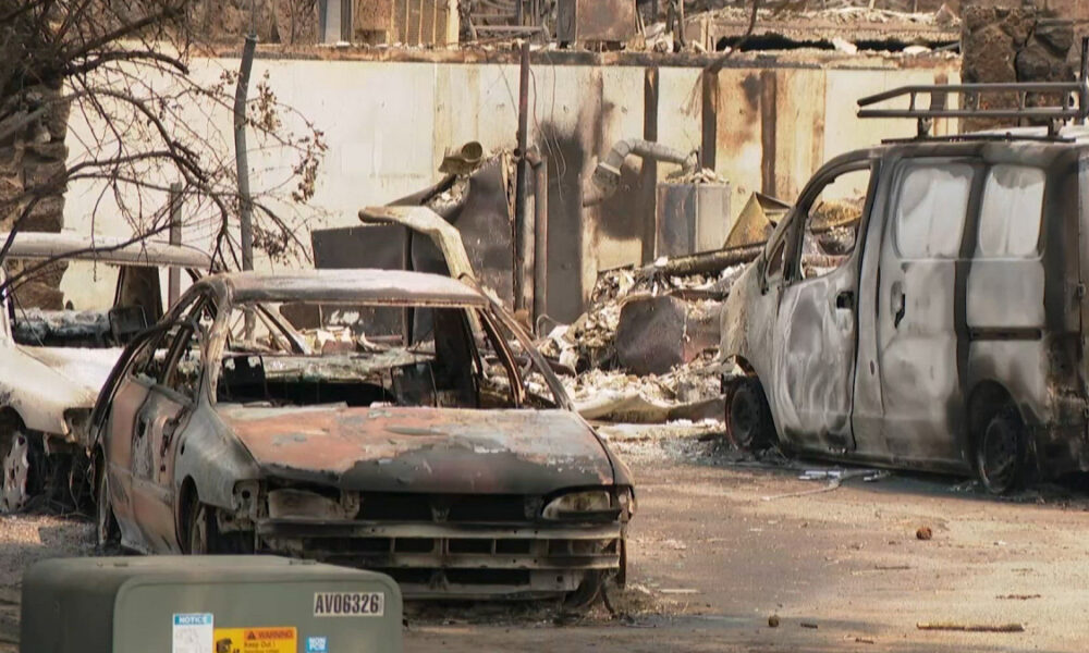 Charred cars and a burned-out van in a debris-strewn lot after a fire, with a ruined wall in the background.