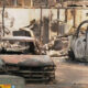 Charred cars and a burned-out van in a debris-strewn lot after a fire, with a ruined wall in the background.