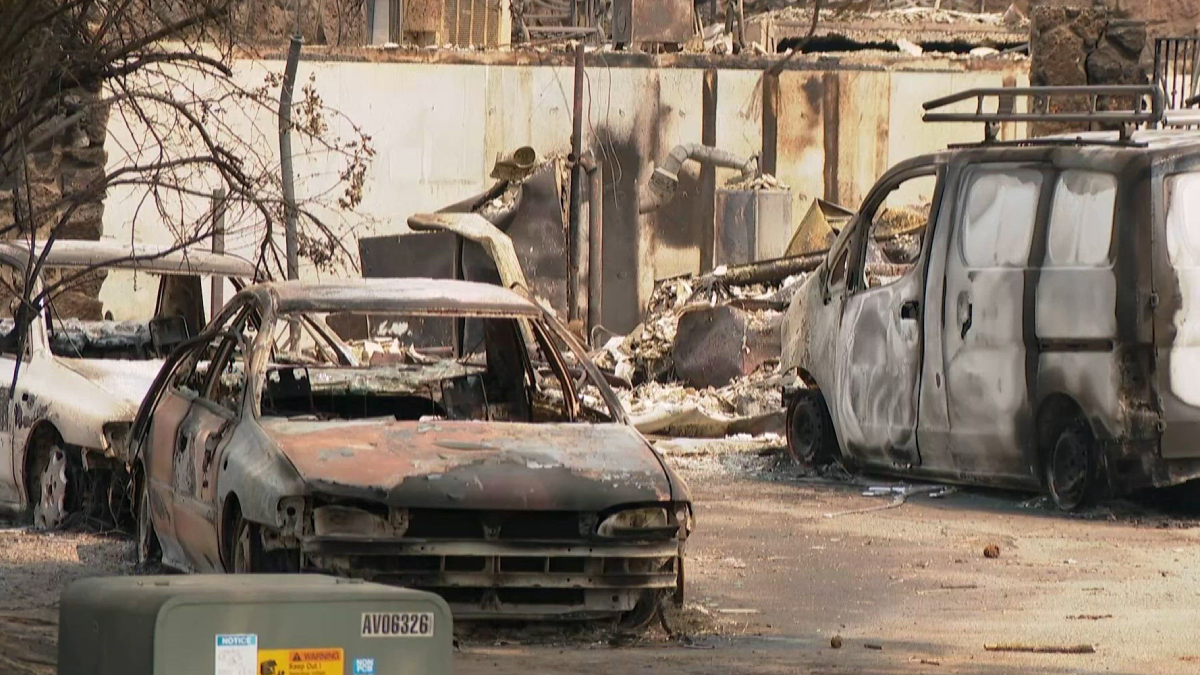 Charred cars and a burned-out van in a debris-strewn lot after a fire, with a ruined wall in the background.