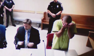 Defendant in a lime-green jail jumpsuit hides his face in a courtroom, with a suited man at the desk and officers in the background.