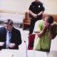 Defendant in a lime-green jail jumpsuit hides his face in a courtroom, with a suited man at the desk and officers in the background.