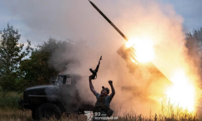 Man in a tactical vest raises a rifle as a rocket launcher detonates a bright flame behind him in a field. The scene shows smoke and a truck in the background.