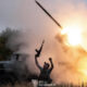 Man in a tactical vest raises a rifle as a rocket launcher detonates a bright flame behind him in a field. The scene shows smoke and a truck in the background.