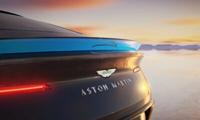 Rear view of a dark Aston Martin with its badge and slim taillight, set against a sunset sky.