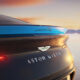 Rear view of a dark Aston Martin with its badge and slim taillight, set against a sunset sky.