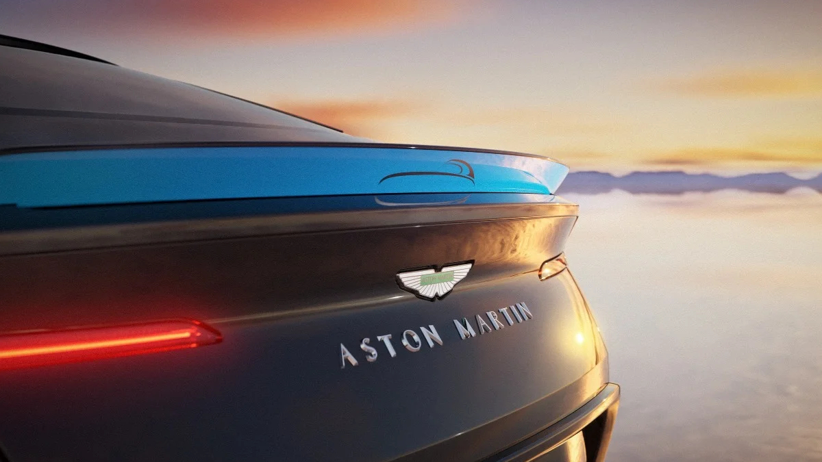 Rear view of a dark Aston Martin with its badge and slim taillight, set against a sunset sky.