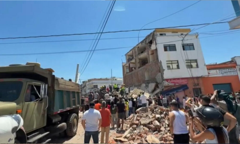 Crowd and rescue workers gather around a collapsed multi-story building with rubble on the street awaiting clearance or rescue efforts.