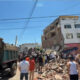 Crowd and rescue workers gather around a collapsed multi-story building with rubble on the street awaiting clearance or rescue efforts.